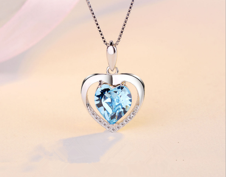 A heart-shaped pendant necklace featuring a faceted light blue heart gemstone in a silver-toned open heart frame with a halo of small clear stones, suspended from a matching silver chain against a soft beige background.