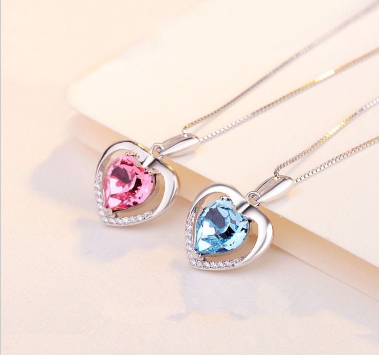 Two silver heart-shaped pendant necklaces with heart-cut gemstones, one pink and one light blue, displayed on a light beige surface.