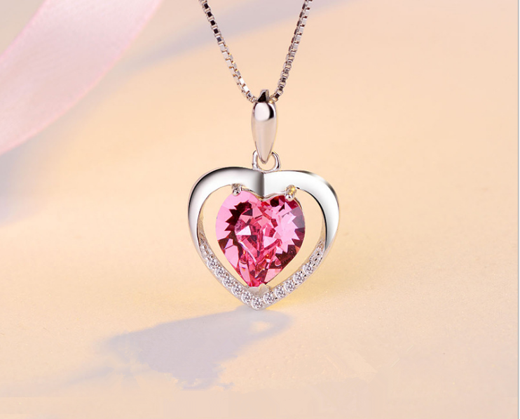 A silver heart-shaped pendant necklace featuring a large faceted pink heart gemstone in the center, with a border of small clear stones, shown on a soft beige background.