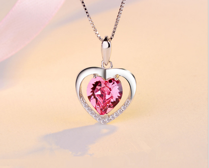 A silver heart-shaped pendant necklace featuring a large faceted pink heart gemstone in the center, with a border of small clear stones, shown on a soft beige background.