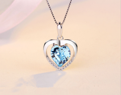 A heart-shaped pendant necklace featuring a faceted light blue heart gemstone in a silver-toned open heart frame with a halo of small clear stones, suspended from a matching silver chain against a soft beige background.